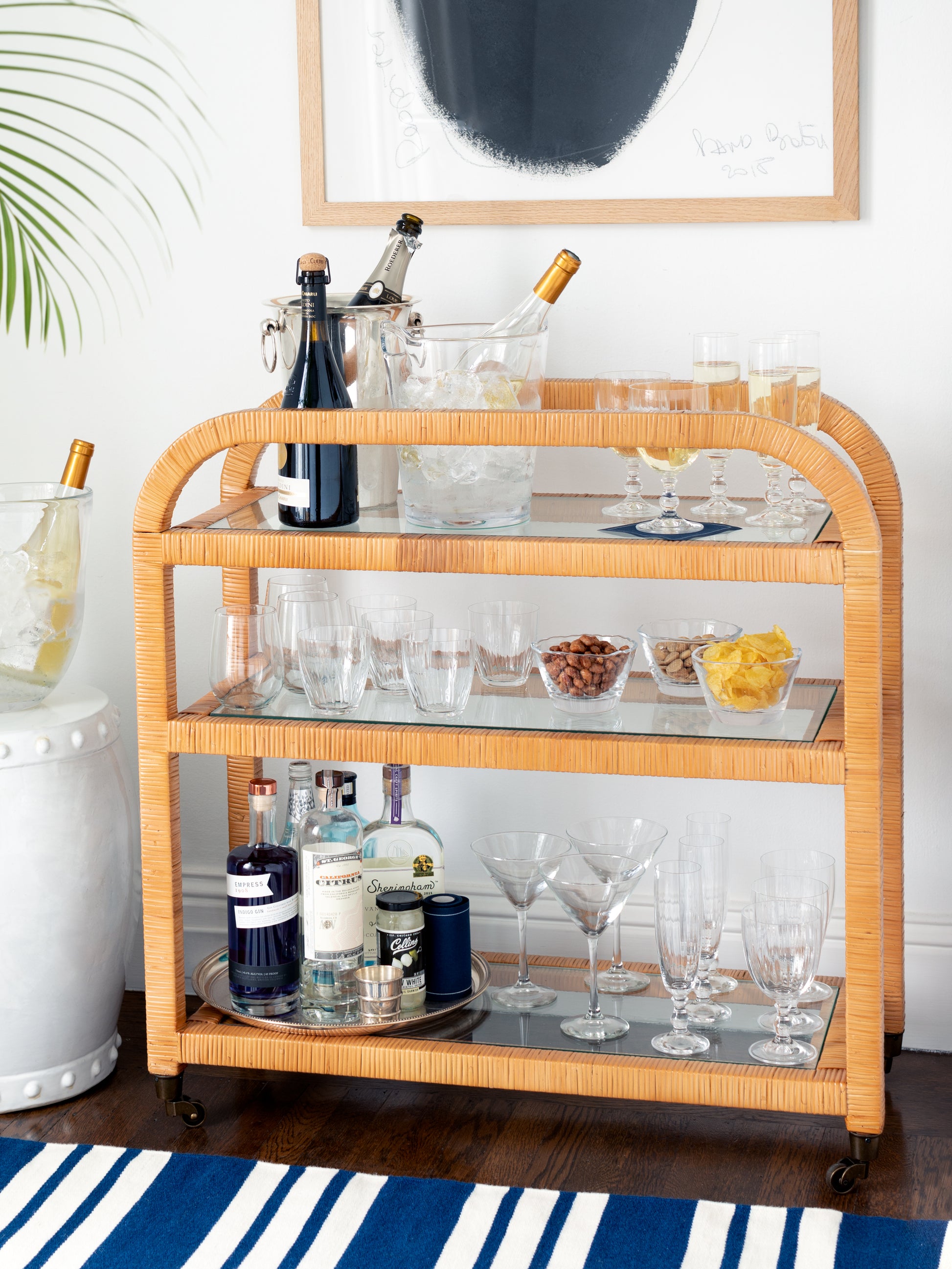Honey rattan Dory Bar Cart with three tiers of shelving, holding drinks, glassware, and snacks.