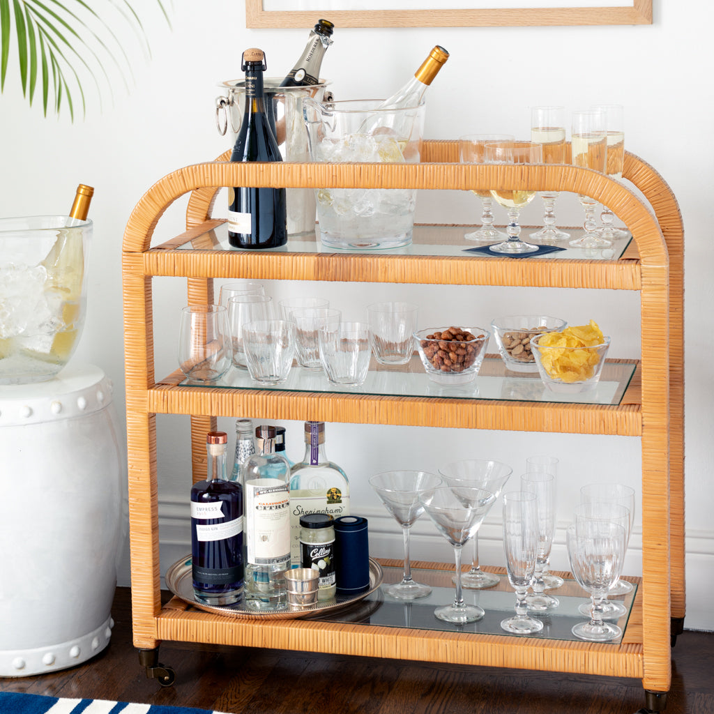 Honey rattan Dory Bar Cart with three glass shelves, holding bottles, glassware, ice buckets, and snacks.