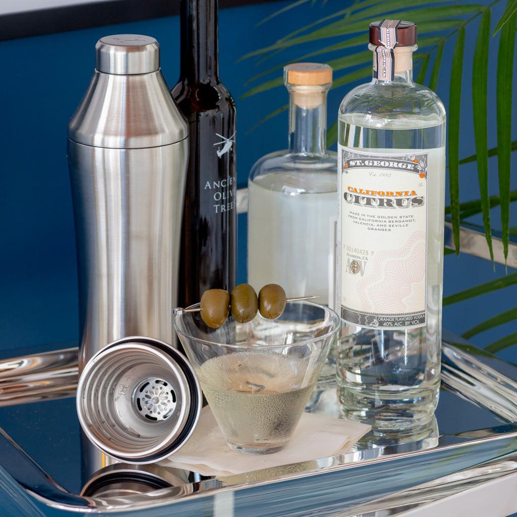 Brushed stainless steel cocktail shaker with integrated measuring top and strainer, martini glass with olives, and bottles on a silver tray.