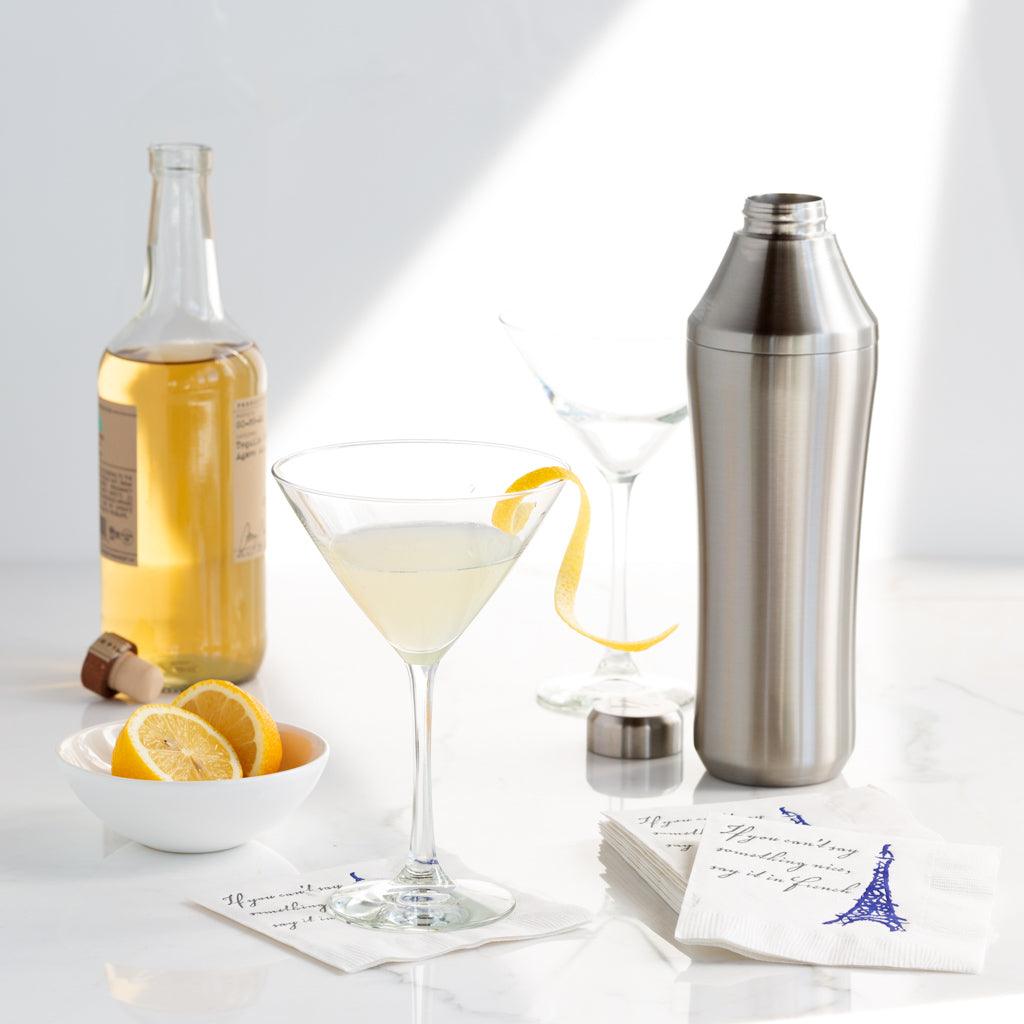 Brushed stainless steel cocktail shaker next to a martini glass with a lemon twist, bottle, and lemons.