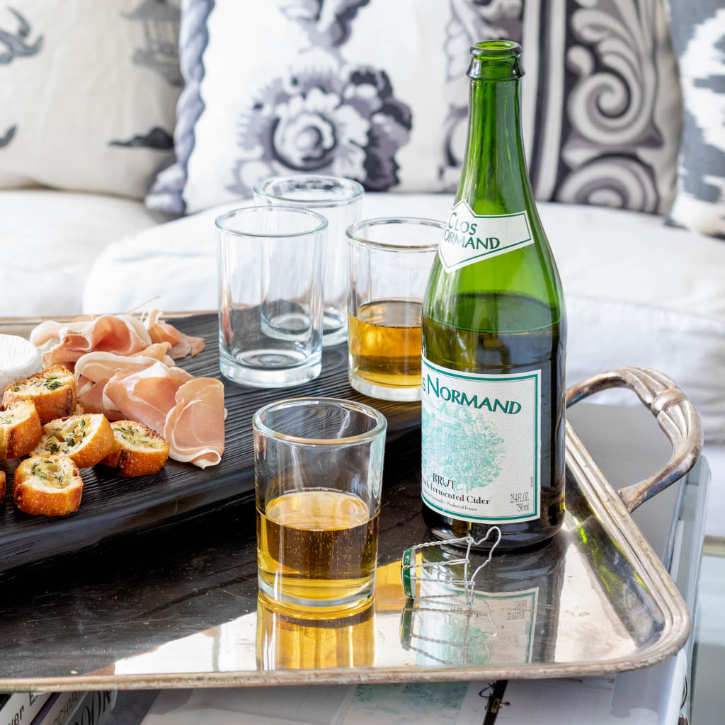 Clear, simple 8.5 oz Everyday Wine Glasses, some filled with amber liquid, next to a green cider bottle and appetizers.