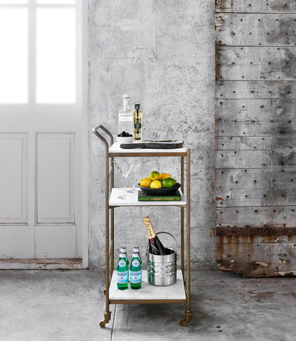 Felix antique brass and white marble square bar cart with three shelves, bottles, and fruit.