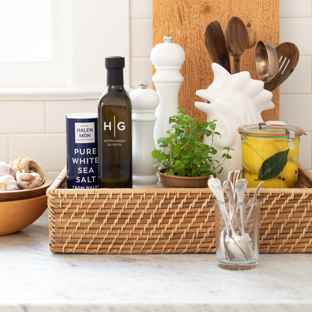 Kitchen counter with a woven tray holding olive oil, sea salt, pepper grinders, herbs, preserved lemons, and silver demitasse spoons.
