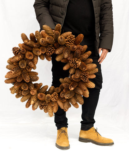 Handmade holiday wreath crafted from natural brown pinecones held by a person against a white background.