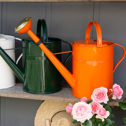 Orange 9-liter zinc watering can with a long spout, next to green and white cans, with pink roses.