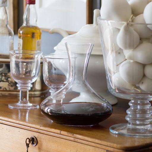 Clear mouth-blown glass wine carafe with a broad base and curved neck, filled with red wine, on a wooden surface.