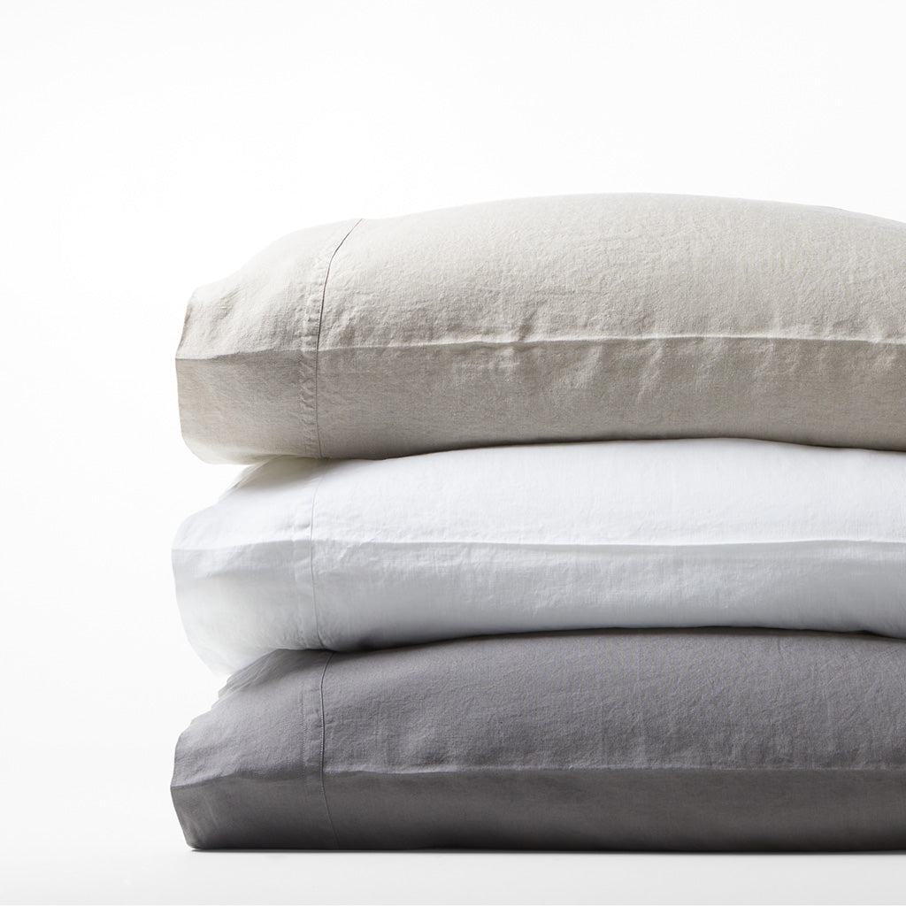 Stack of three Washed-Linen pillowcases: light grey, white, and dark grey, with double-needle stitching.