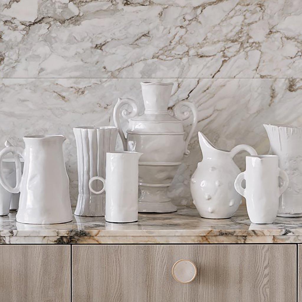 Handmade creamy white ceramic bumpy pitcher 5163 by Montes Doggett, surrounded by other white ceramic vessels on a marble counter.
