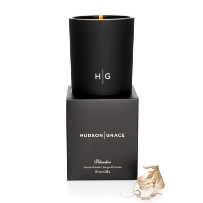 Hudson Grace Bleecker Scented Candle in a matte black glass vessel with a burning wick, atop its black gift box.