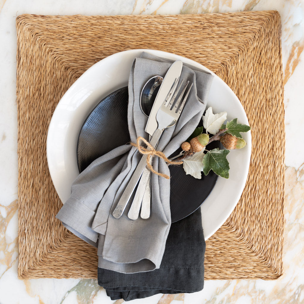 Natural abaca woven square placemat with white and black plates, grey napkin, and cutlery.