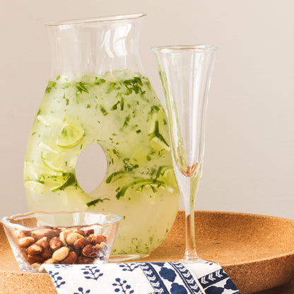 Clear, thick, air-blown glass champagne flute with a rippled rim, next to a pitcher of lime drink and nuts.