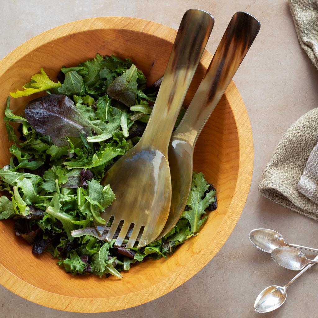 Oversized light horn salad servers with natural grain in a wooden bowl of greens.