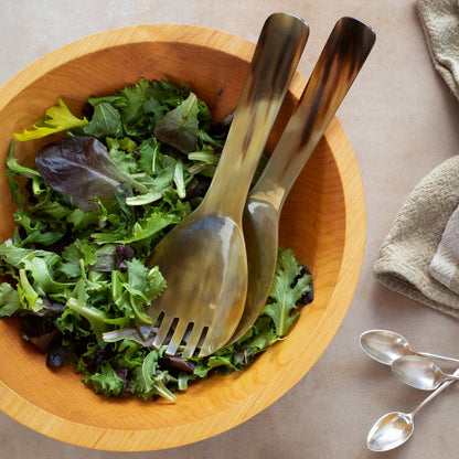 Oversized light horn salad servers with natural grain in a wooden bowl of greens.