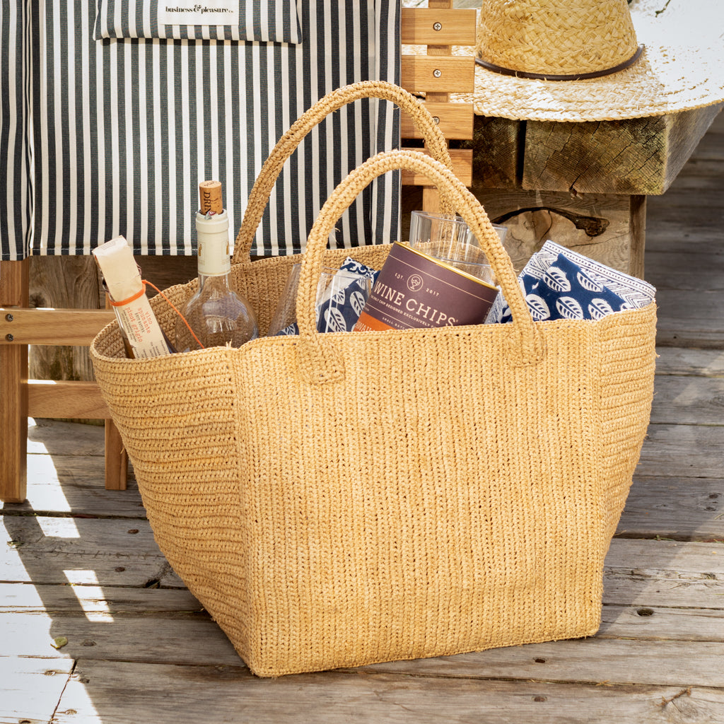 Handcrafted, packable raffia tote bag in natural tan, filled with picnic items.