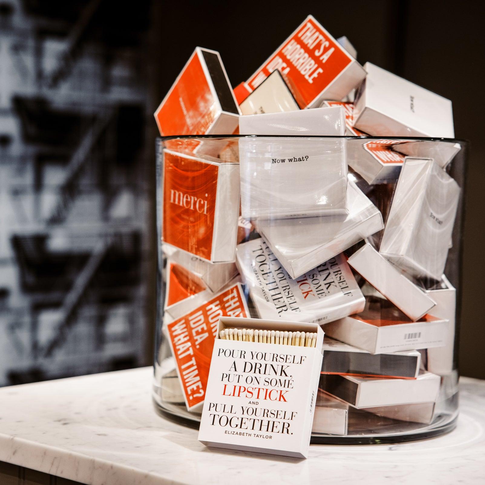 Oversized "Merci" matches in a clear glass jar, with one box open showing long white matchsticks.