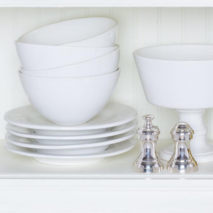 Stack of white ceramic bowls and plates with two silver shakers on a white shelf.