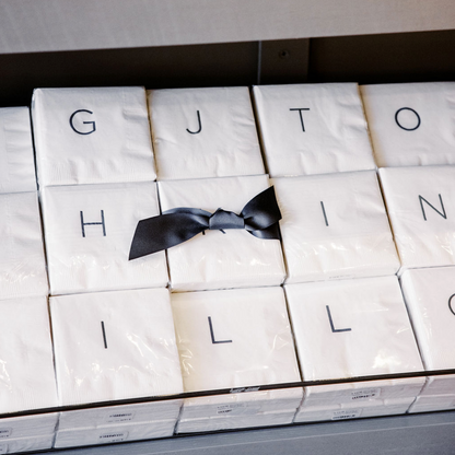 White 3-ply paper cocktail napkins with grey sans-serif monograms, tied with a black bow.