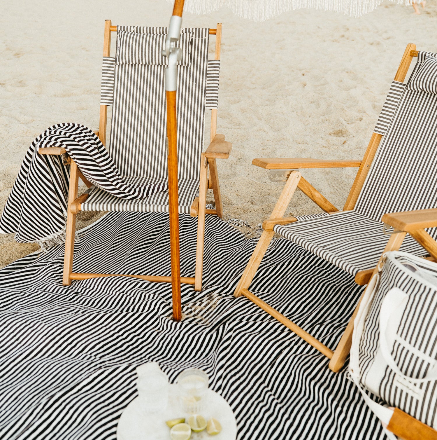 Navy and white stripe canvas reclining beach chairs with teak frames, a matching blanket, and cooler bag on sand.