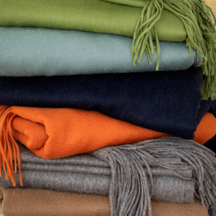 Stack of cashmere throw blankets in navy blue, green, light blue, orange, gray, and tan with fringe.