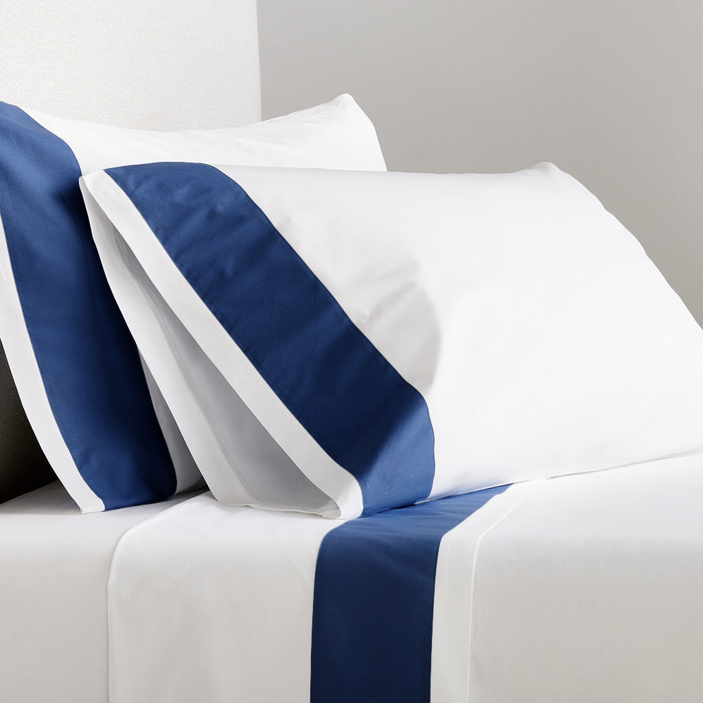 Two white percale pillowcases with a navy blue wide-band detail, on a bed with matching sheets.