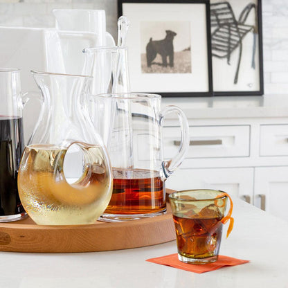 Clear glass "O" pitcher with white wine, alongside other pitchers and a drink in a green glass.