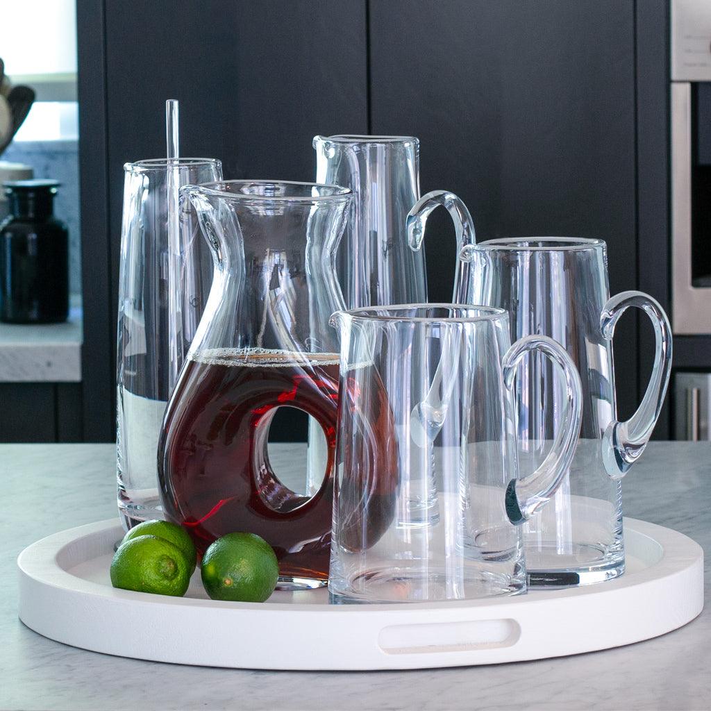 Clear "O" glass pitcher with iced tea, surrounded by various clear glass pitchers and tumblers on a white tray with limes.