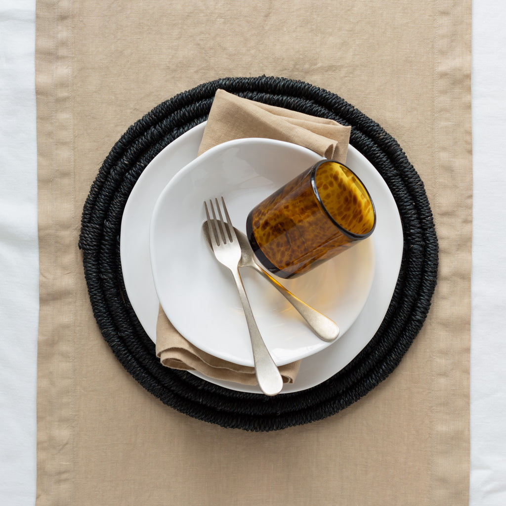 Organic Low Bowl, creamy white ceramic, 9" diameter, on a black woven placemat with a tan napkin and glass.