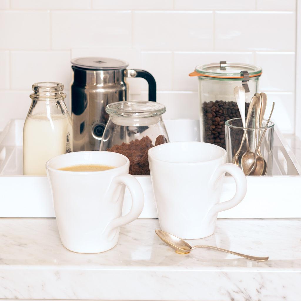 Two white ceramic mugs with organic edges, one filled with coffee, on a marble counter with coffee accessories.