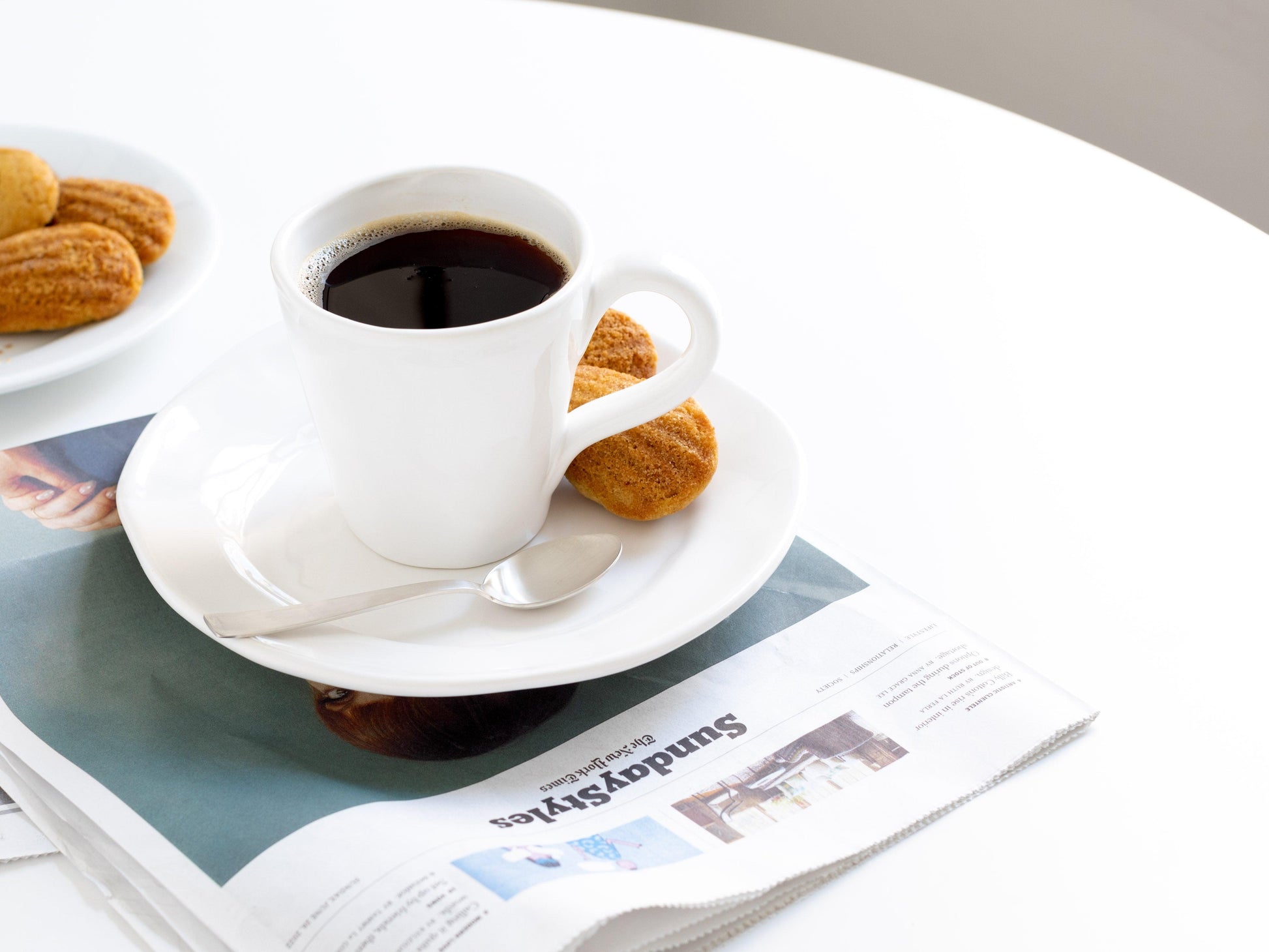 Organic white ceramic mug with coffee, saucer, spoon, and madeleines on a newspaper.