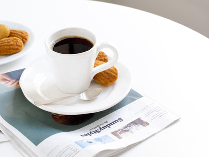 Organic white ceramic mug with coffee, saucer, spoon, and madeleines on a newspaper.