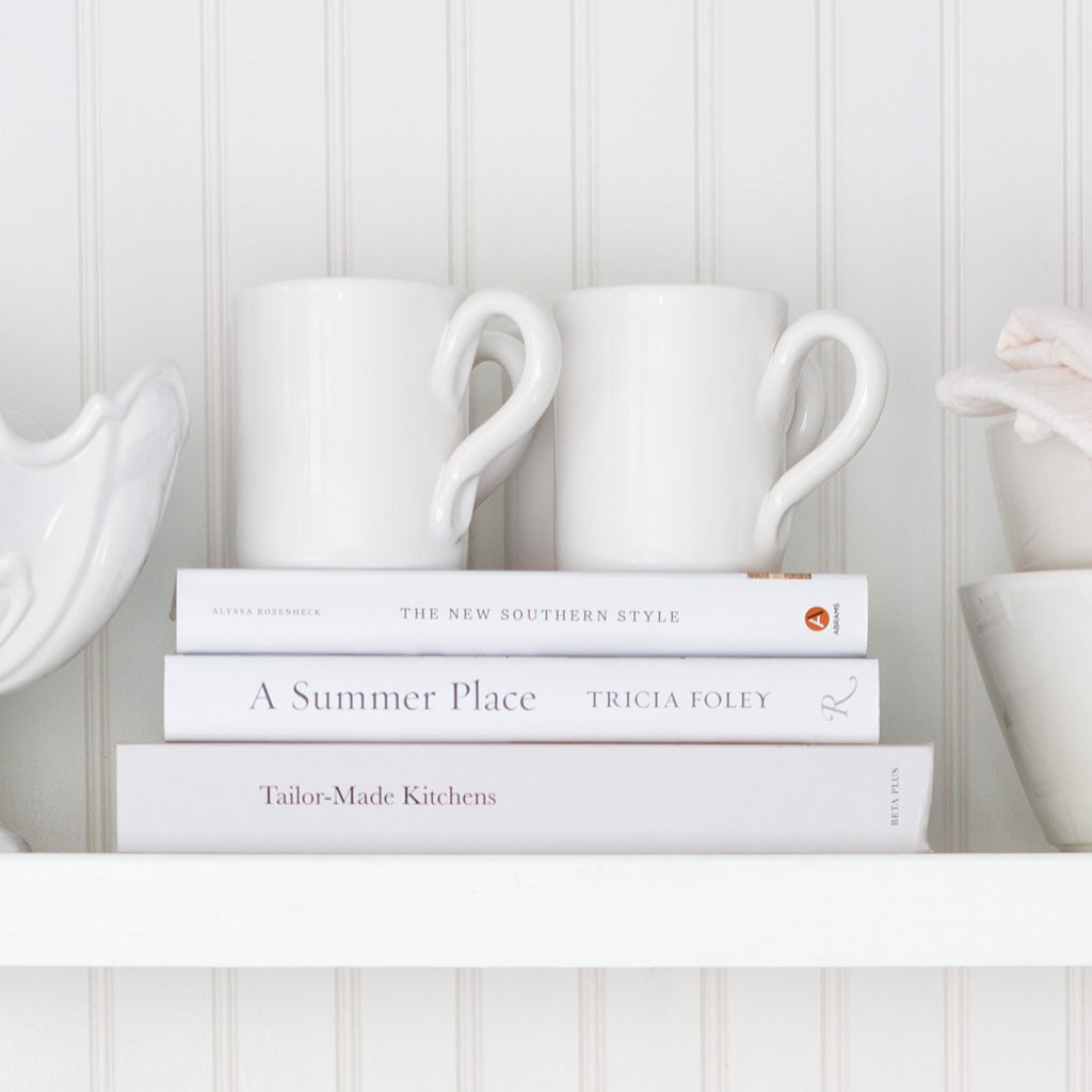 Two creamy white HG Original ceramic mugs with clean edges and curved handles, stacked on books.