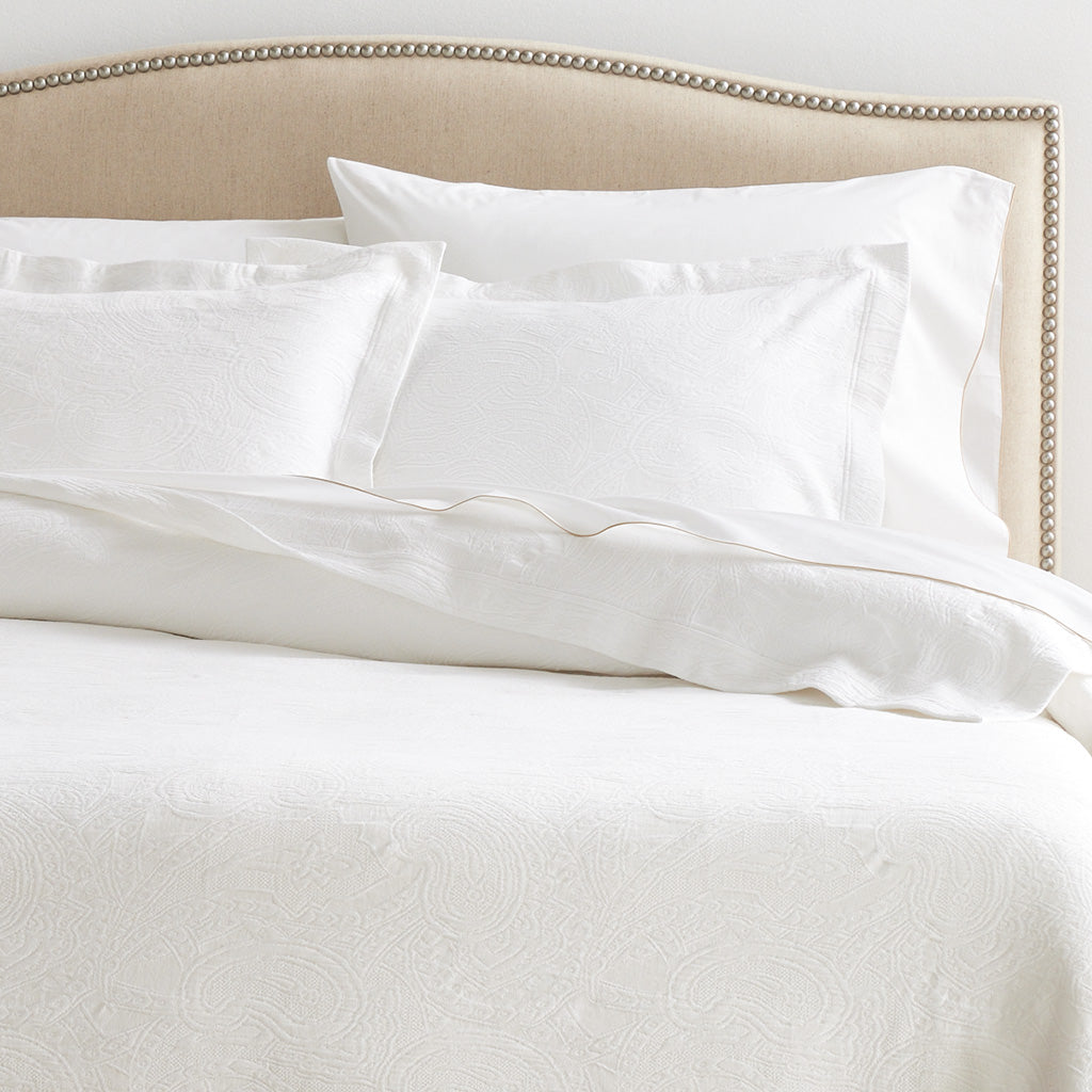 White cotton matelassé coverlet and shams with subtle paisley pattern on a bed with a beige headboard.