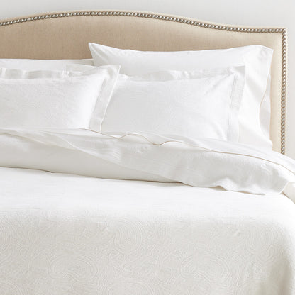 White cotton matelassé coverlet and shams with subtle paisley pattern on a bed with a beige headboard.