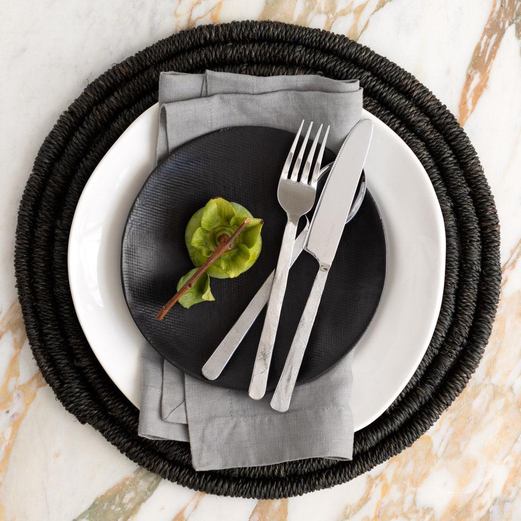 Rain HG Signature hand-dyed light grey linen napkin, 22-inch, folded on a black plate with cutlery.