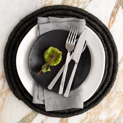 Rain HG Signature hand-dyed light grey linen napkin, 22-inch, folded on a black plate with cutlery.