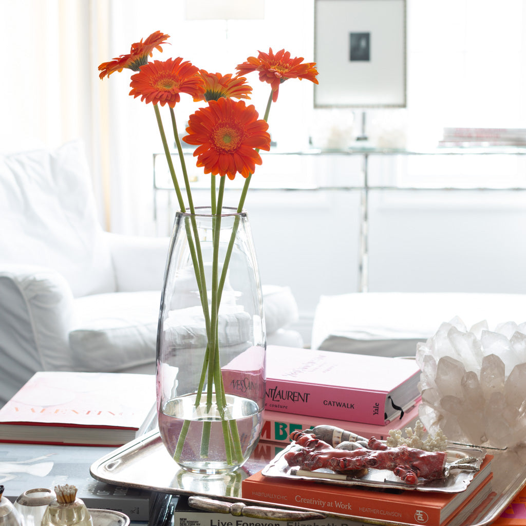 Tall, clear mouth-blown glass Robert Vase with orange gerbera daisies on a silver tray.