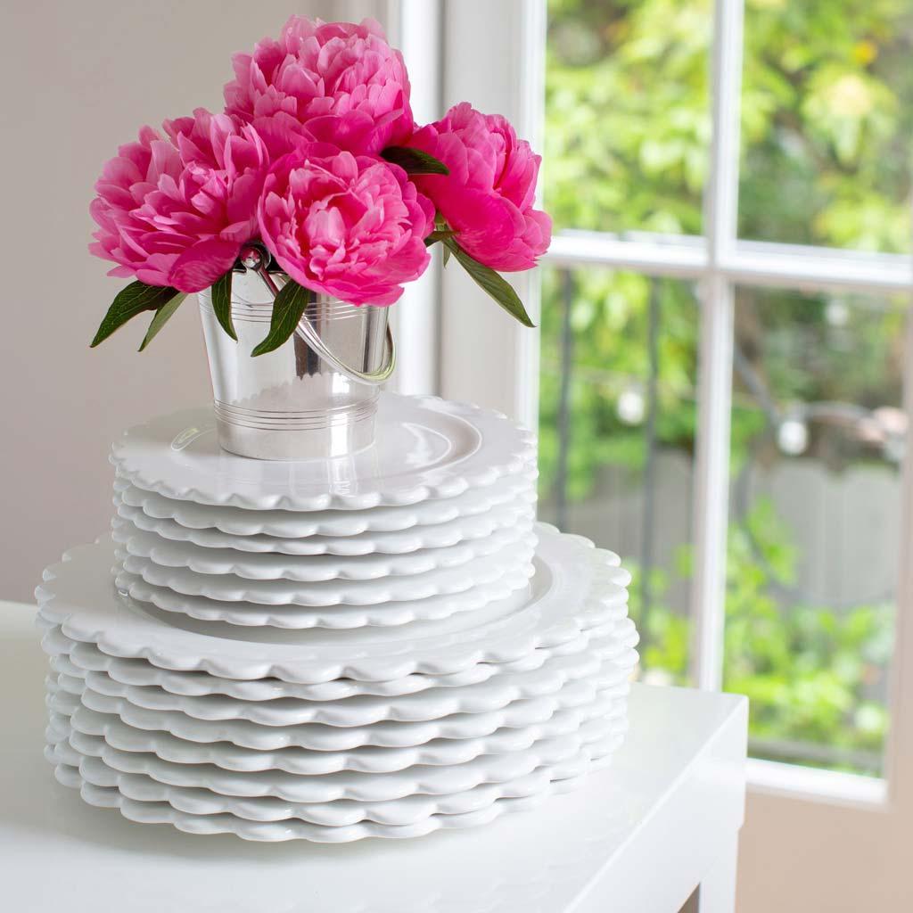 Stack of white ceramic scalloped dinner plates with a small bucket of pink peonies.