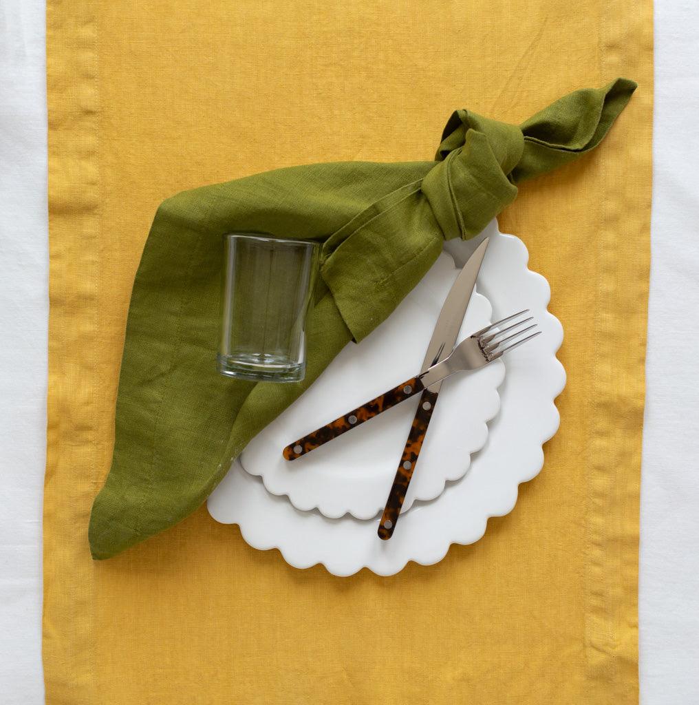 White ceramic scalloped dinner plate with salad plate, green linen napkin, clear glass, and tortoise-handle flatware.