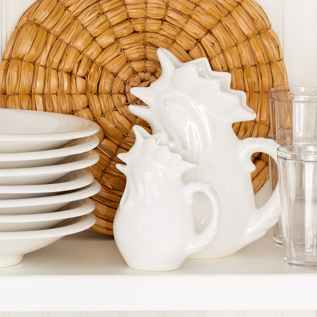 Two white ceramic rooster pitchers, one small and one large, on a shelf with white plates and glasses.