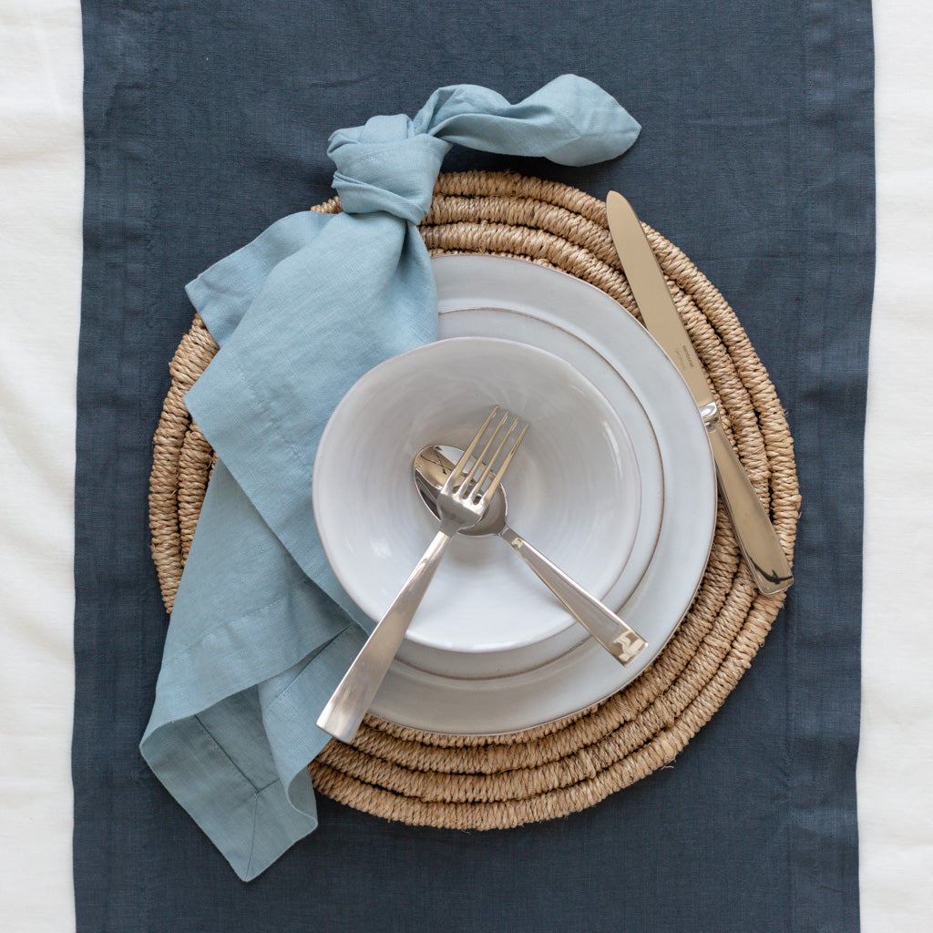 Round natural woven abaca placemat, 15" Dia, with light blue napkin, white plates, and silverware.