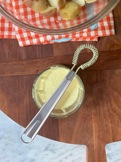 Stainless steel mini whisk with a coiled head resting in a glass of yellow sauce.
