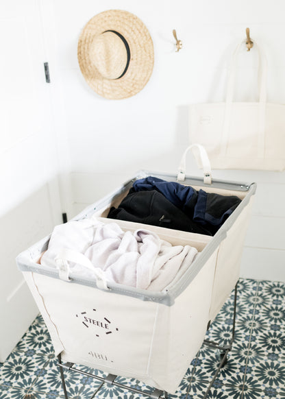 Steele 4-bushel canvas elevated rolling laundry hamper with steel frame, filled with clothes, on a patterned floor.
