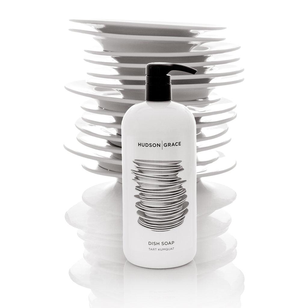 White Hudson Grace Tart Kumquat Dish Soap bottle with black pump, in front of stacked white plates.