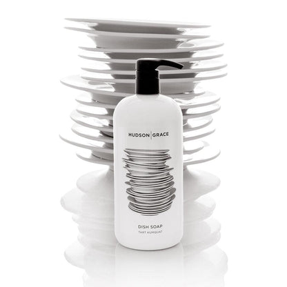 White Hudson Grace Tart Kumquat Dish Soap bottle with black pump, in front of stacked white plates.