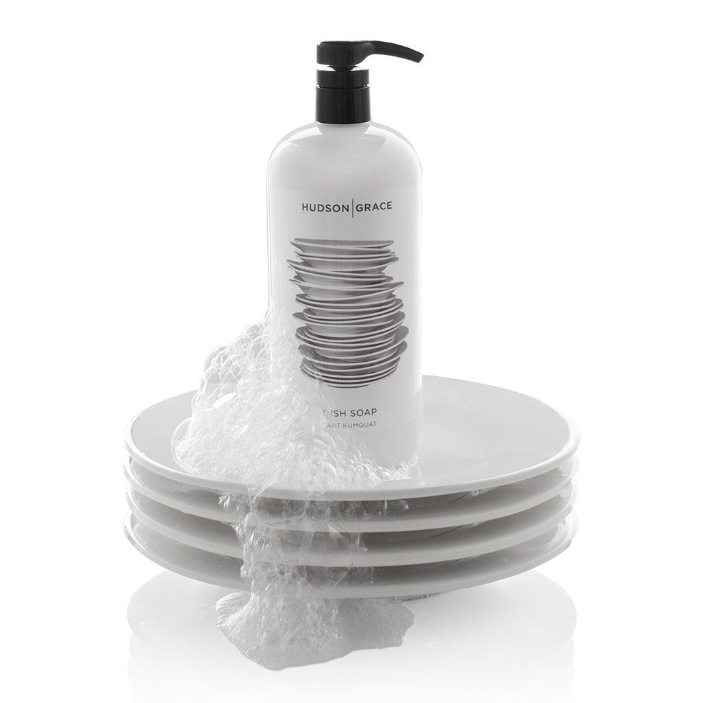 White HG Tart Kumquat Dish Soap bottle with black pump, 32 oz, on a stack of white plates with suds.