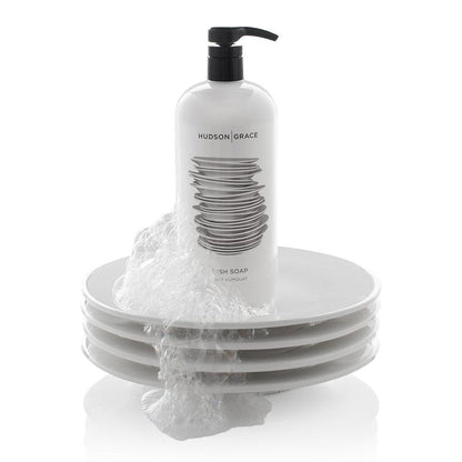 White HG Tart Kumquat Dish Soap bottle with black pump, 32 oz, on a stack of white plates with suds.