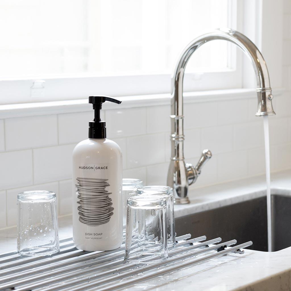 White HG Tart Kumquat Dish Soap bottle with black pump, on a drying rack with clear glasses by a sink.
