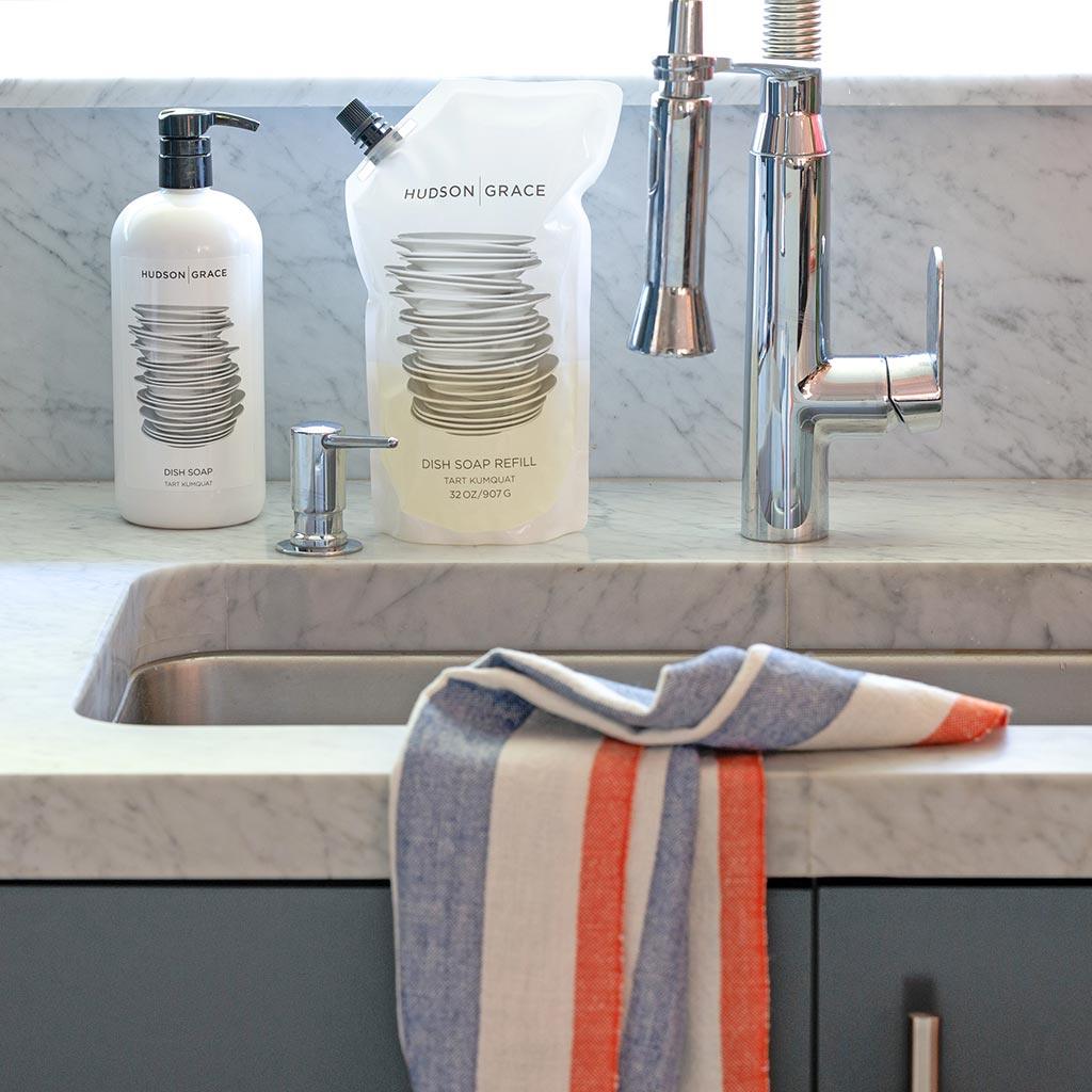 White HG Tart Kumquat Dish Soap bottle, refill pouch, and chrome dispenser on a marble counter.