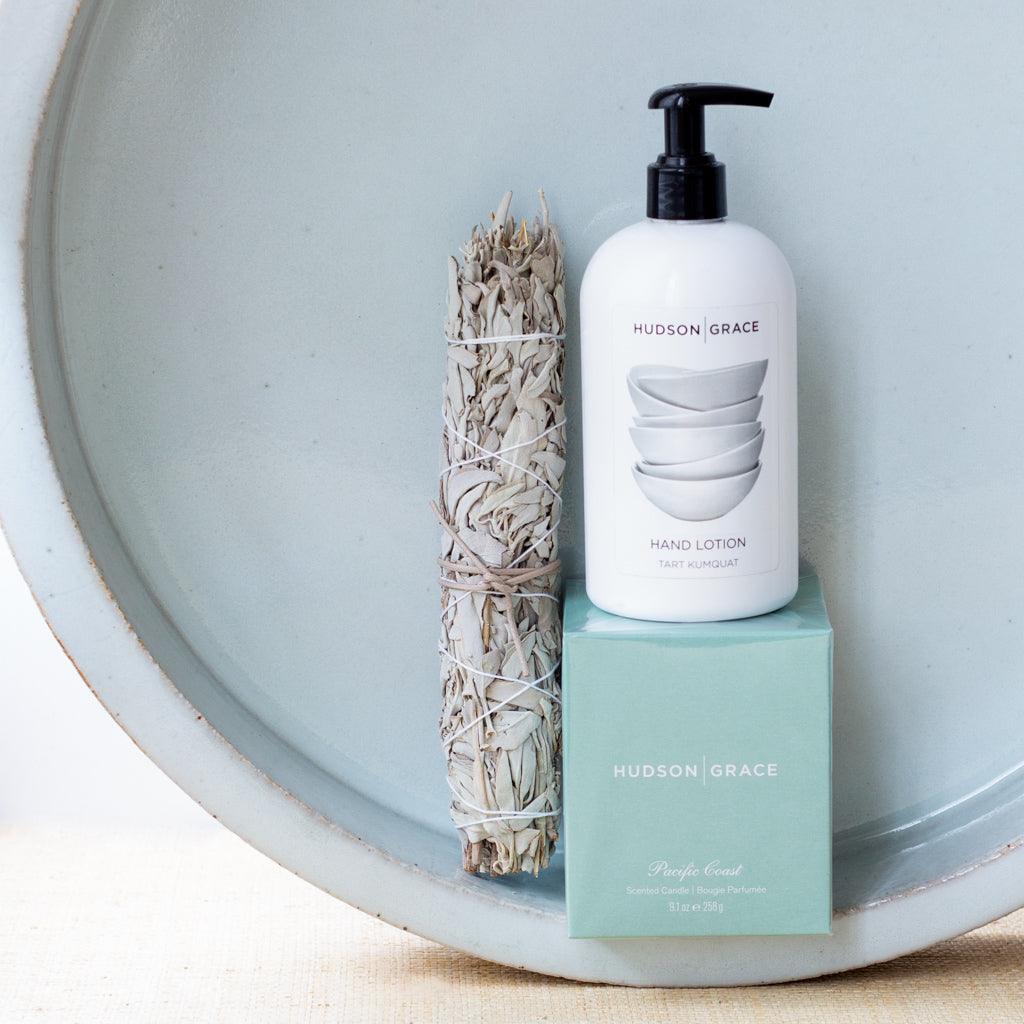 White Hudson Grace Tart Kumquat Hand Lotion bottle with black pump, sage bundle, and teal candle box on a light blue tray.