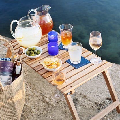 Teak portable outdoor picnic table with drinks, snacks, and a woven bag by the water.